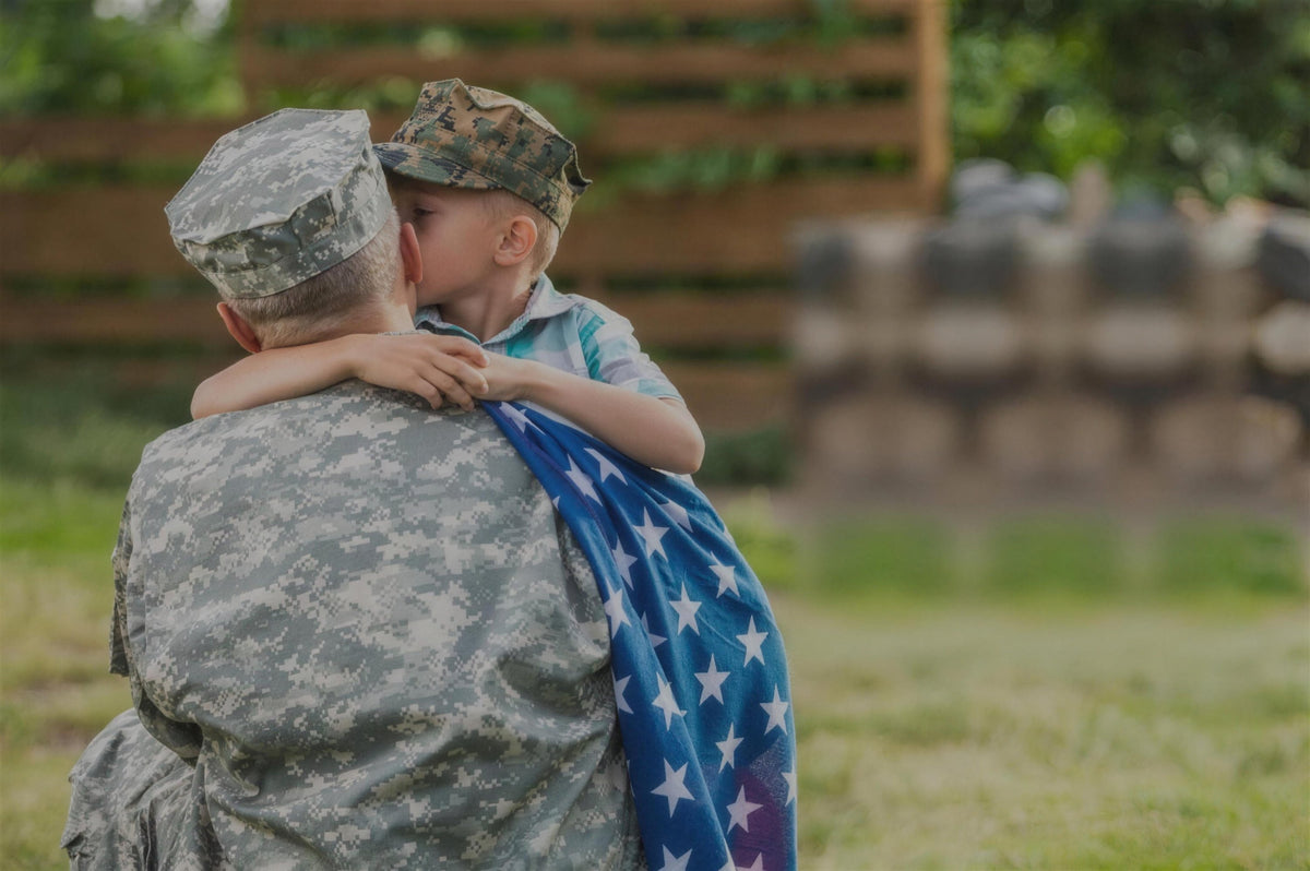A young boy in a camo cap kisses his father, a soldier in uniform with an American flag over his shoulder